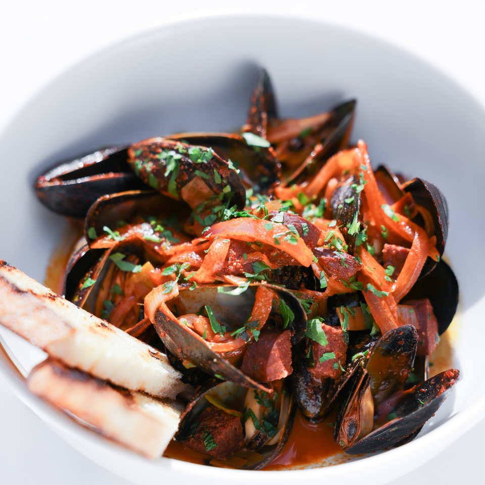 Mussels in a rich red sauce with chorizo, onions, and parsley, served with grilled bread.