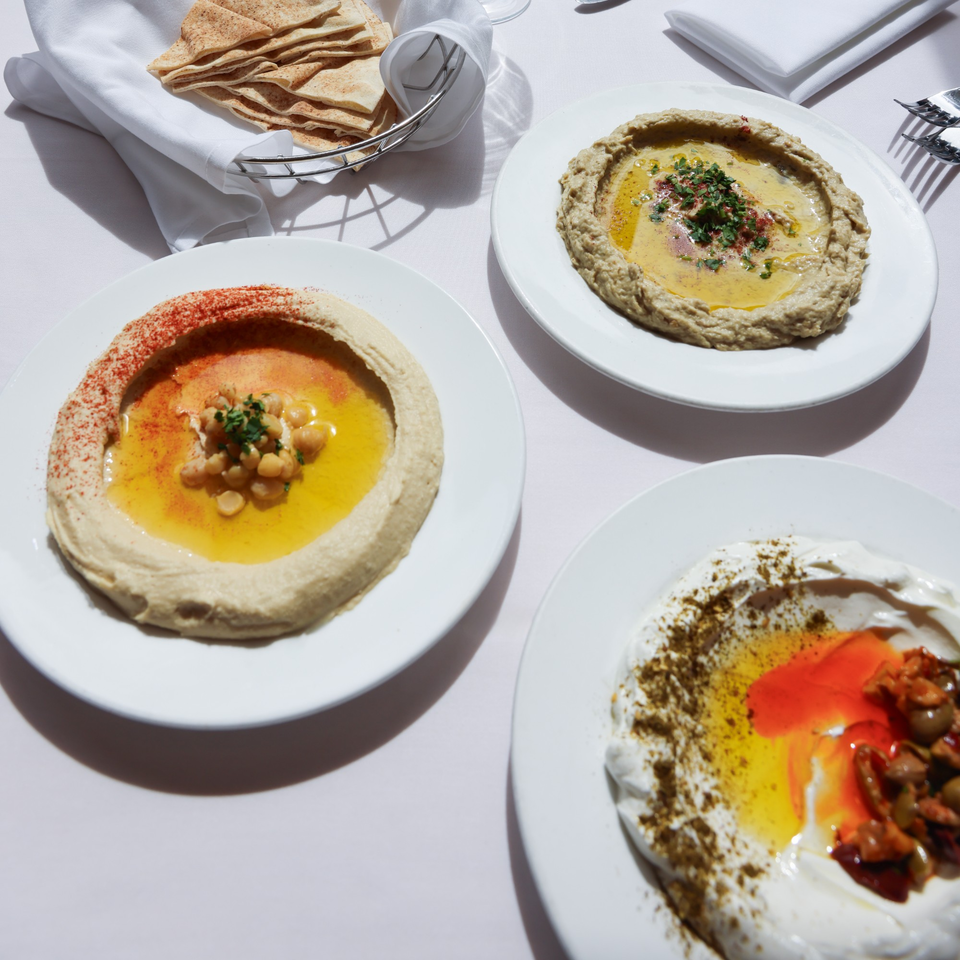 Three Mediterranean dips, including hummus and baba ghanoush, served with pita bread on a white tablecloth.